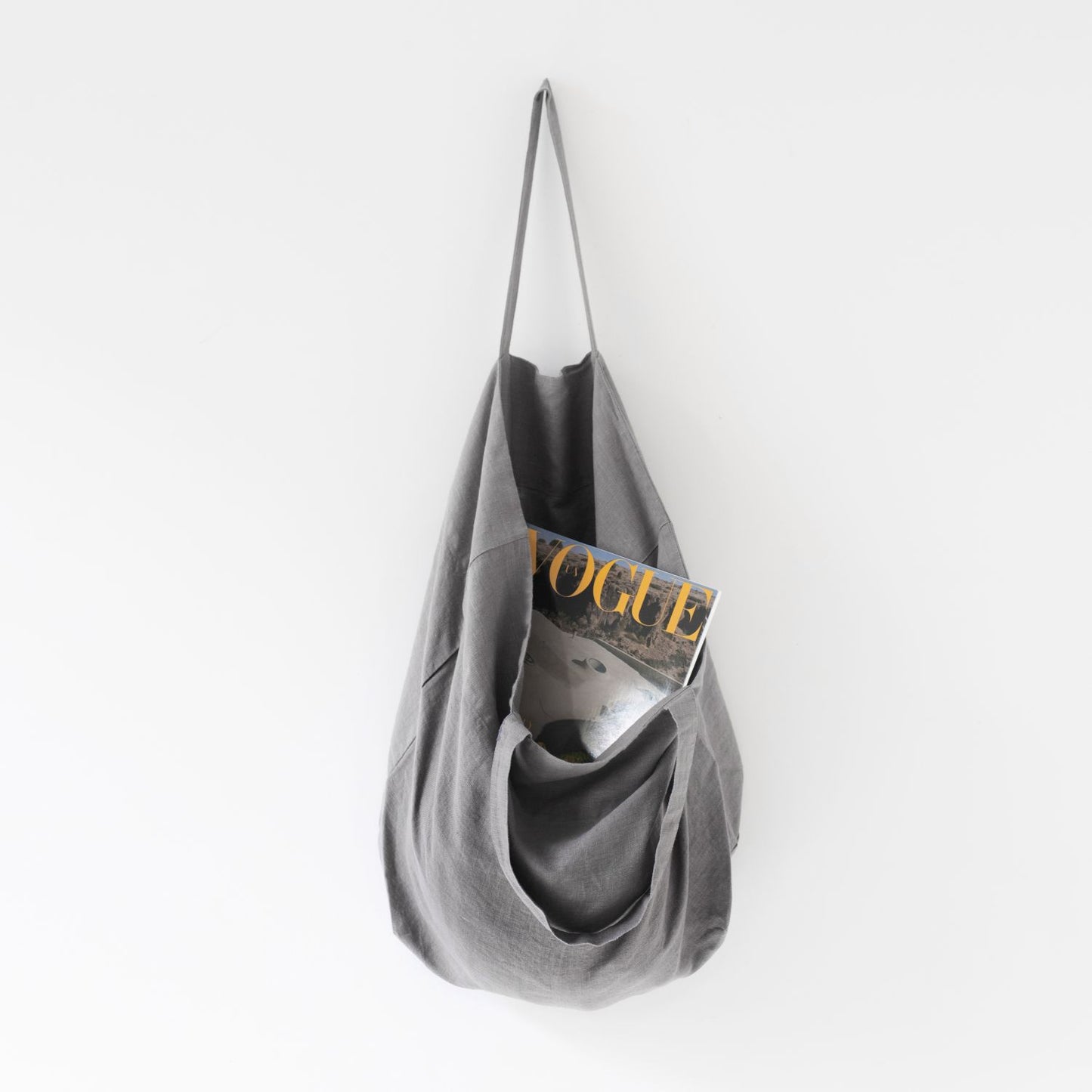 Medelstor tygväska i grå linne som hänger på en krok med sitt ena handtag. Väskan har formen av en rektangel med medellånga handtag. Från väskan skymtar en Vouge tidning.