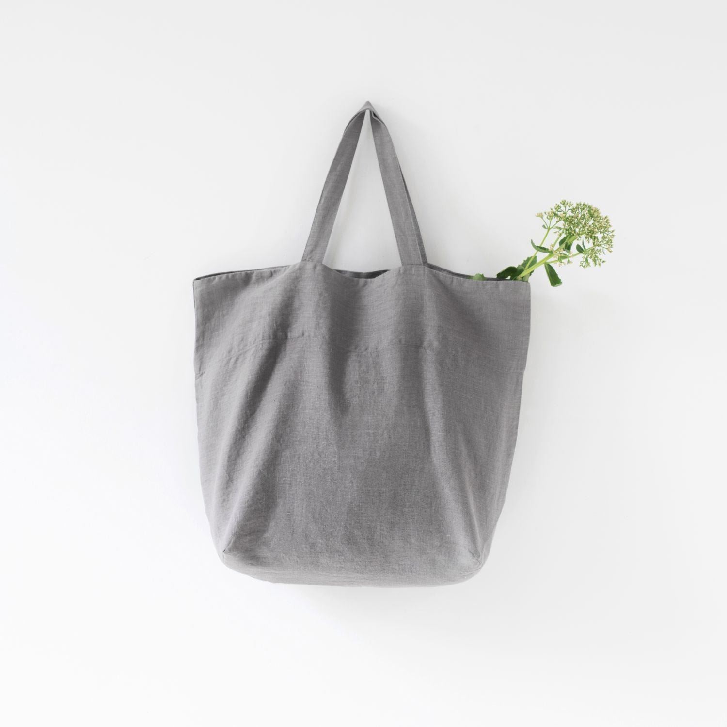 Medelstor tygväska i ljusgrå linne som hänger på en krok. Väskan har formen av en rektangel med medellånga handtag. Från väskan sticker en grön kvist upp.