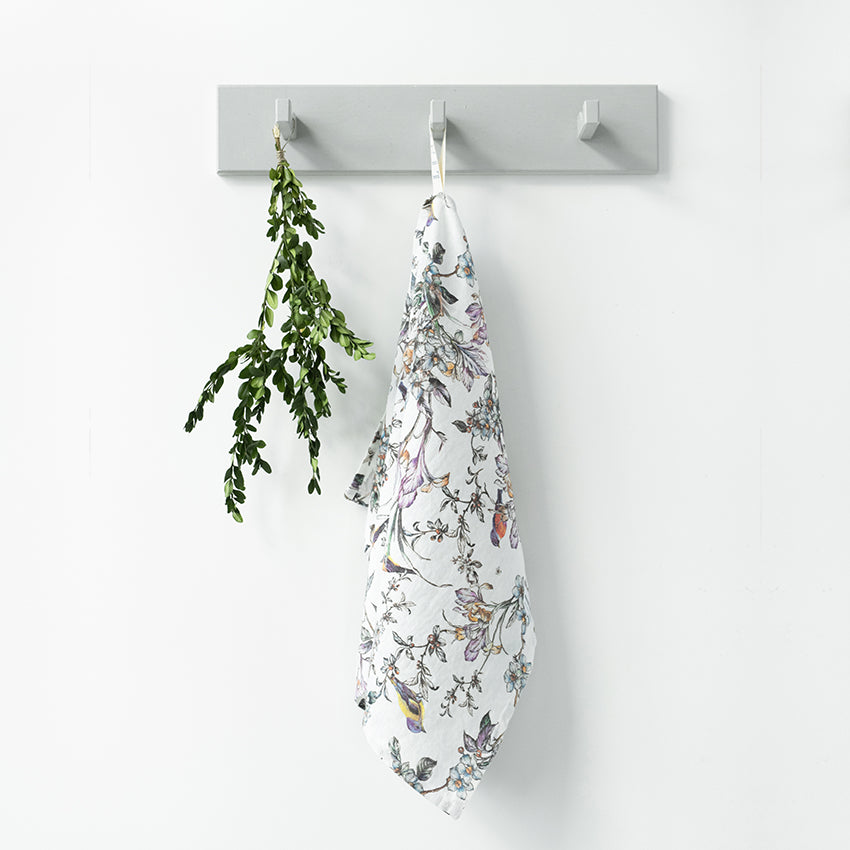 En kökshandduk i mönstrat linne. Mönstret har fåglar och blommor i olika dova färger och hänger på en grå knopplist med sin vita ögla. 