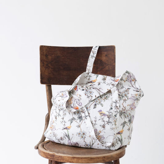 Tygväska i linne med medellånga handtag och mönster med fåglar och blommor i ljusa färger står på en stol. 