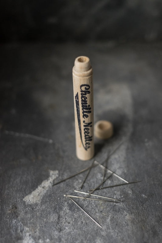 Tio små silvriga broderinålar ligger utslängda på en stenskiva och bredvid står förpackningen i trä.