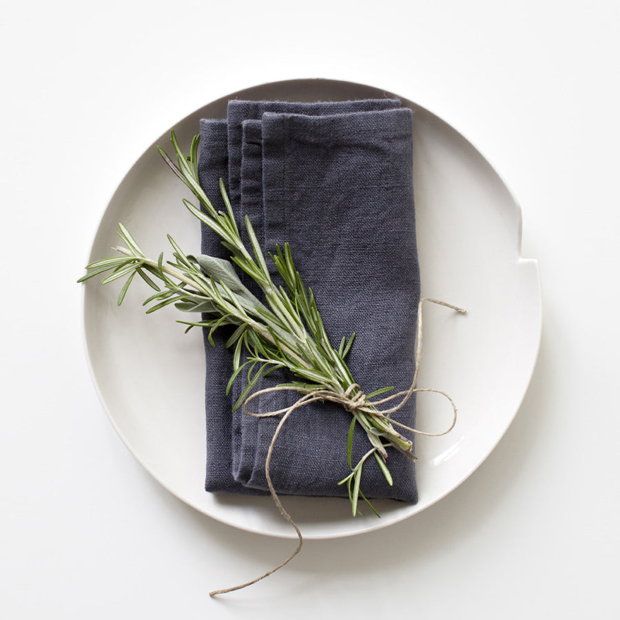 En handsydd tygservett i mörkgrå linne som ligger vikt på en vit tallrik tillsammans med kvistar av timjan.