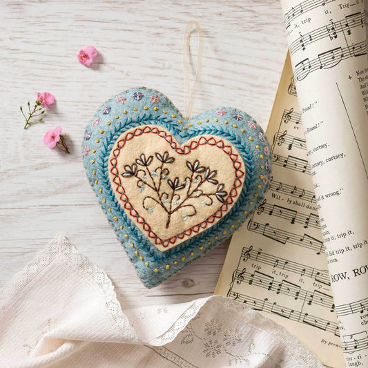 Ett blått hjärta med små detaljer på i filt ligger på ett träbord. En vitt band sitter på hjärtat för upphängning. 