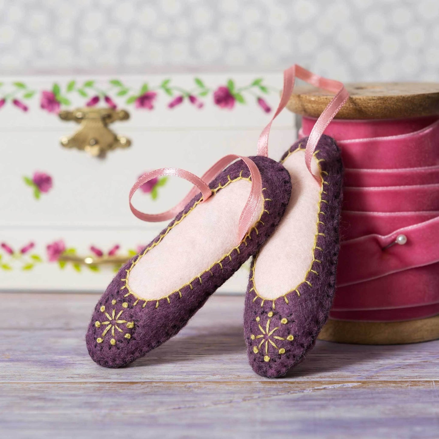 Ett par lila ballerinaskor i filt med ljusrosa sidenband för upphängning. Skorna står lutandes mot en ceriserosa rulle av sammetsband. 