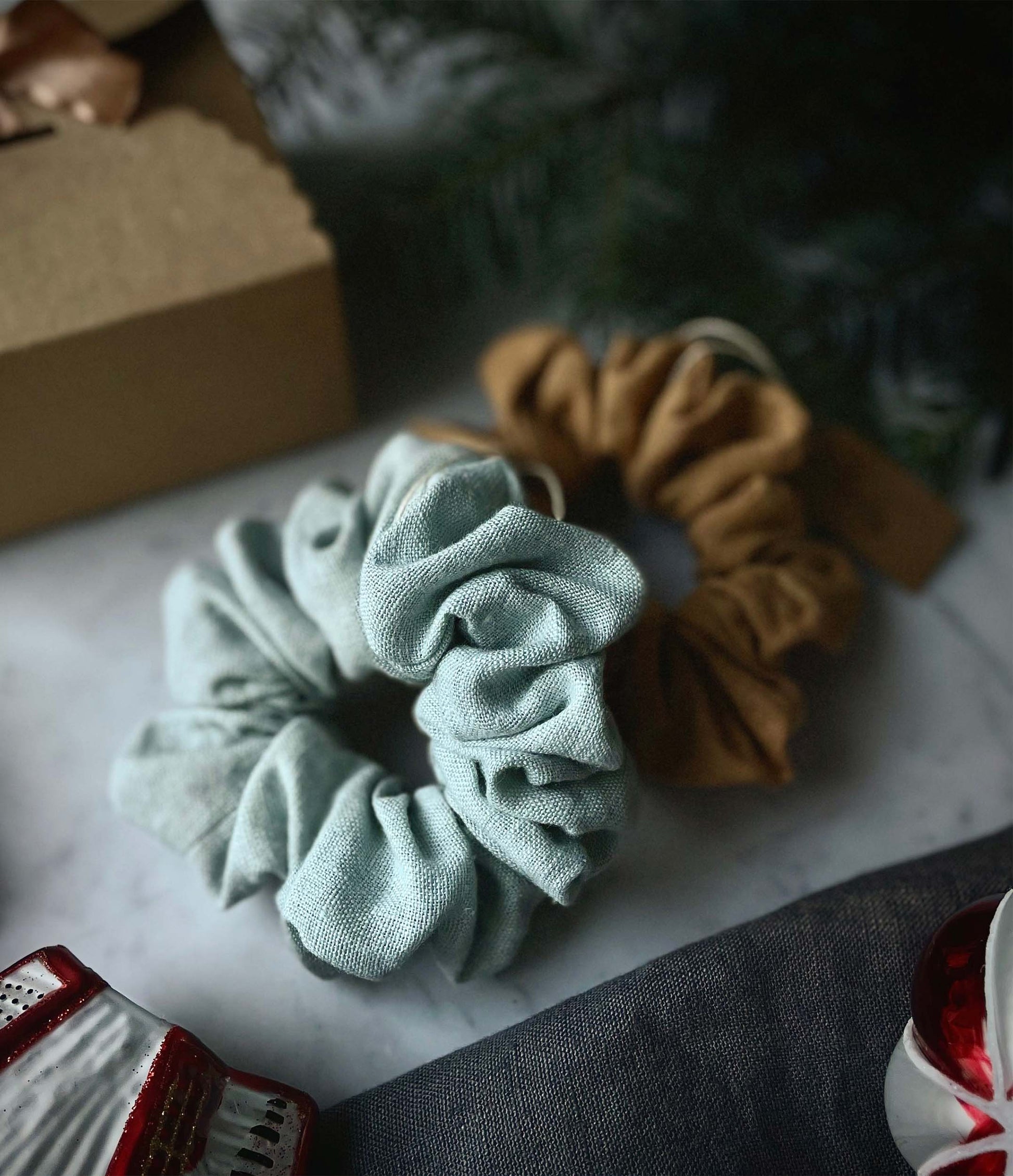 Två handgjorda linne scrunchies i blå och rosfärg ligger på ett bord tillsammans med en miljövänlig papperskartong.