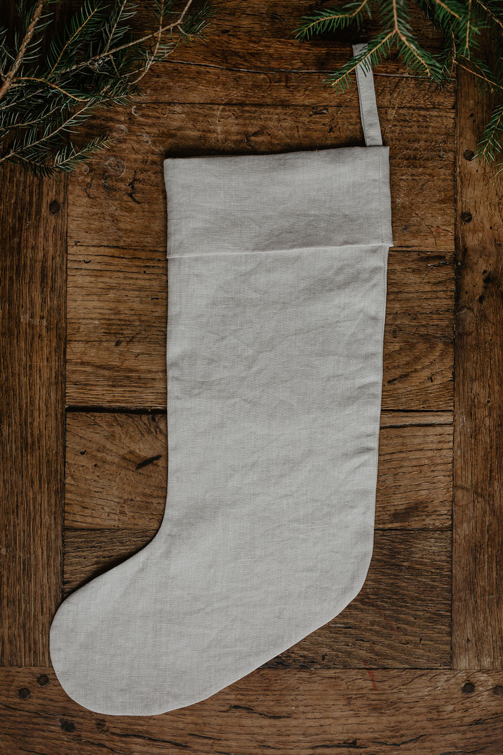 En vit julstrumpa i linne med nedvikt kant och ögla ligger på en träskiva tillsammans med granris.