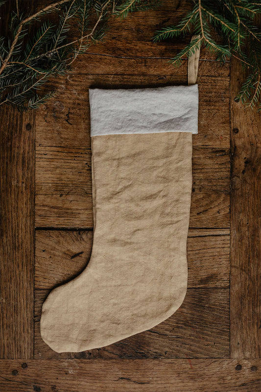 En handsydd beige julstrumpa i linne med ögla som ligger på ett träbord med granris.
