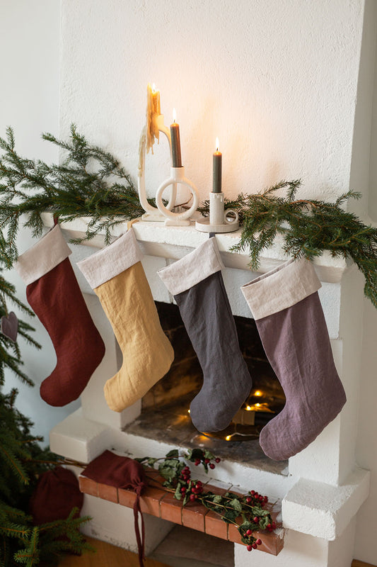 Fyra stycken julstrumpor i linne med olika färger hänger framför en eldstad inomhus.