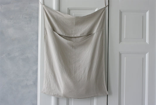En stor tvättpåse i naturellt linne som hänger på en dörr. En öppningsficka skymtar på framsidan av påsen. 