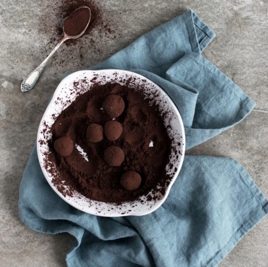 En blå kökshandduk i linne ligger skrynkligt på ett grått bord. På handduken står ett spillkum med innehåll av kakao och chokladtryfflar. I bakgrunden skymtar en tesked fylld med kakaopulver.