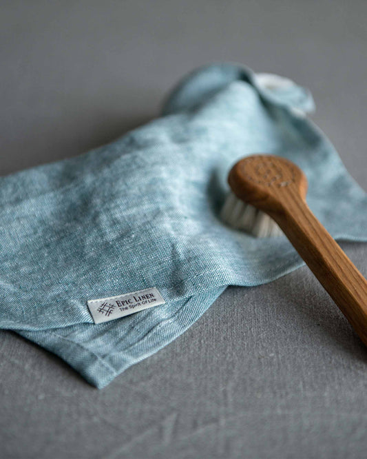 Tvättlapp i blå linnefärg som ligger på en grå linneduk. Varumärkets namn är fastsytt på långsidan där det står Epic Linen. 