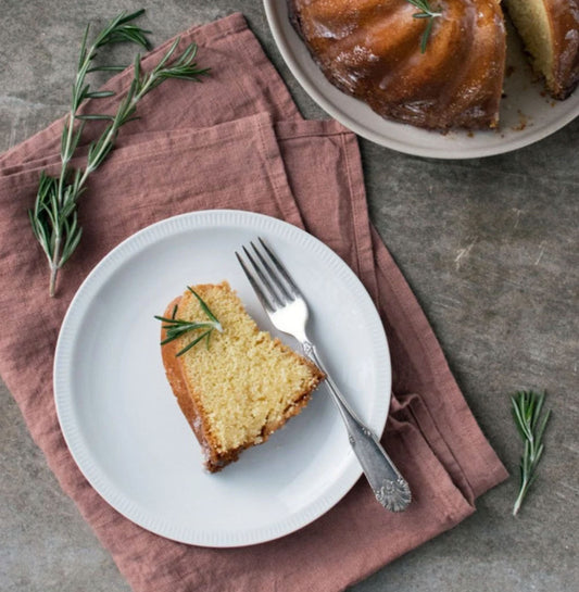 En kökshandduk i aprikosfärgad linne ligger vikt på ett stenbord och på servetten står det en vit desserttallrik med en sockerkaka på. 