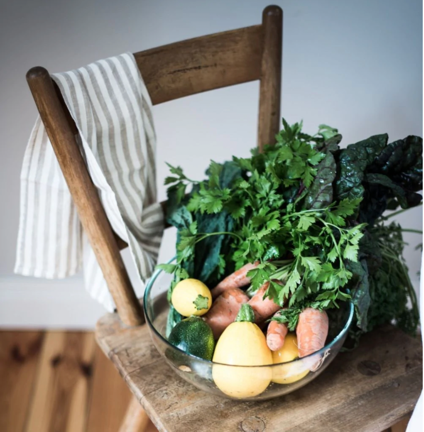 En kökshandduk i vit och beiget randigt linne hänger över en stolskant. På trästolen står en medelstor glasskål fylld med olika sorters grönsaker och kryddor.