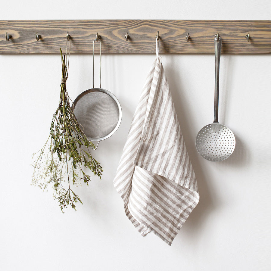 En handsydd kökshandduk i vit och beige randigt linne som hänger med sin ögla på en knopplist i trä.