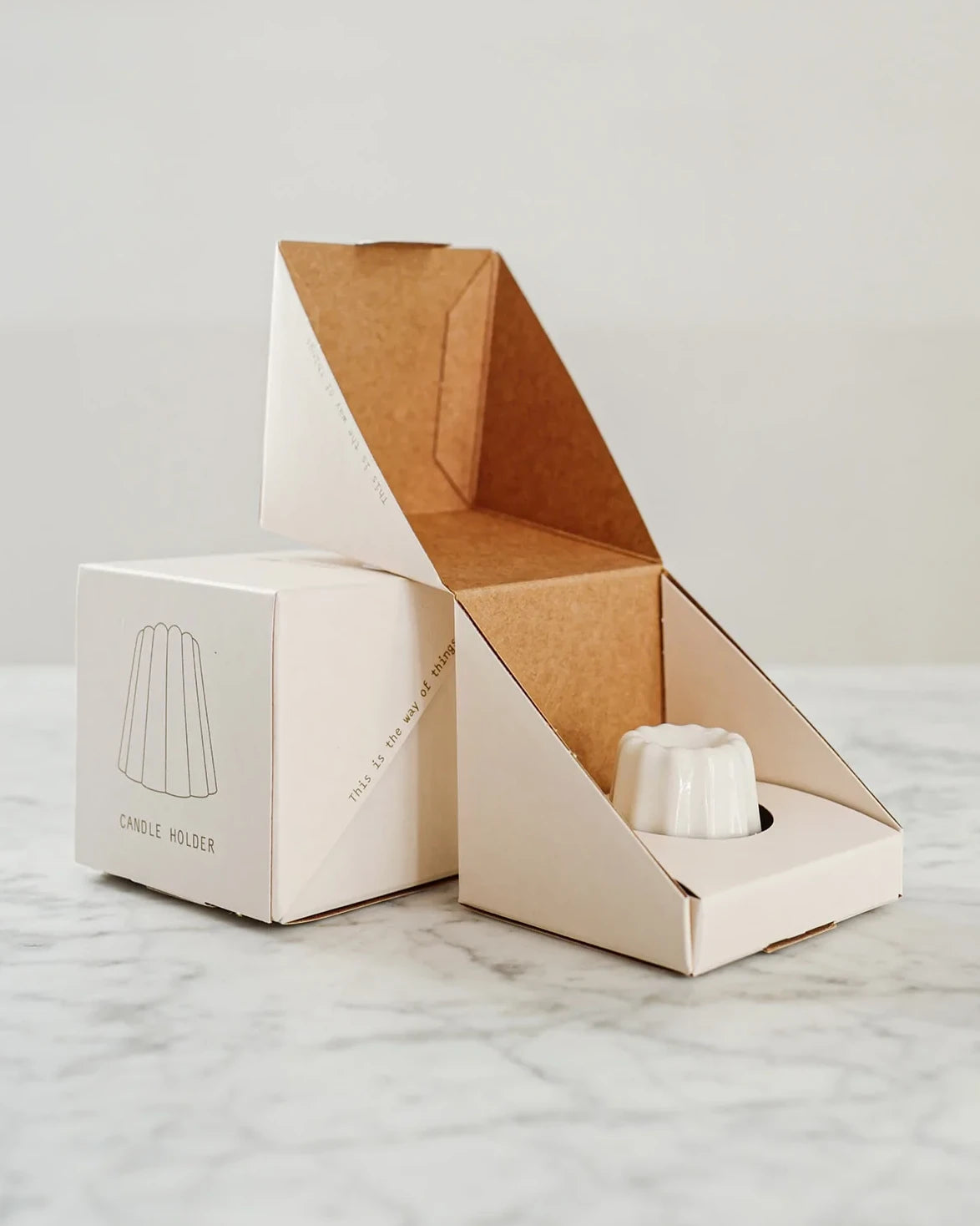 En liten handgjord ljushållare i vit keramik ligger i sin vita pappersförpackning. 