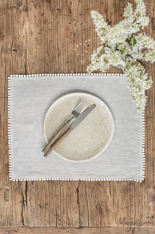 Rektangulär ljusgrå bordtablett i linne med en kantdetalj i form av små söta tygbollar. Bordtabletten ligger på ett rustikt träbord med en vit blomma. 