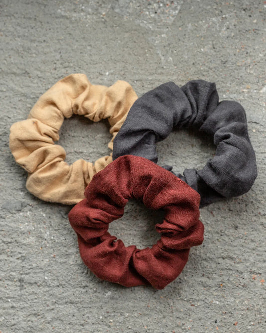Tre stycken scrunchies i linne i olika färger ligger bredvid varandra på en grå yta. 