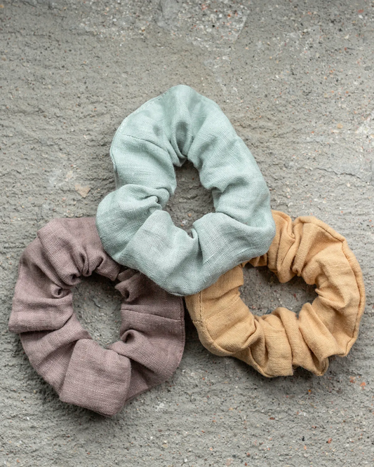 Tre stycken scrunchies i dova färger i materialet lin ligger bredvid varandra på en grå yta. 