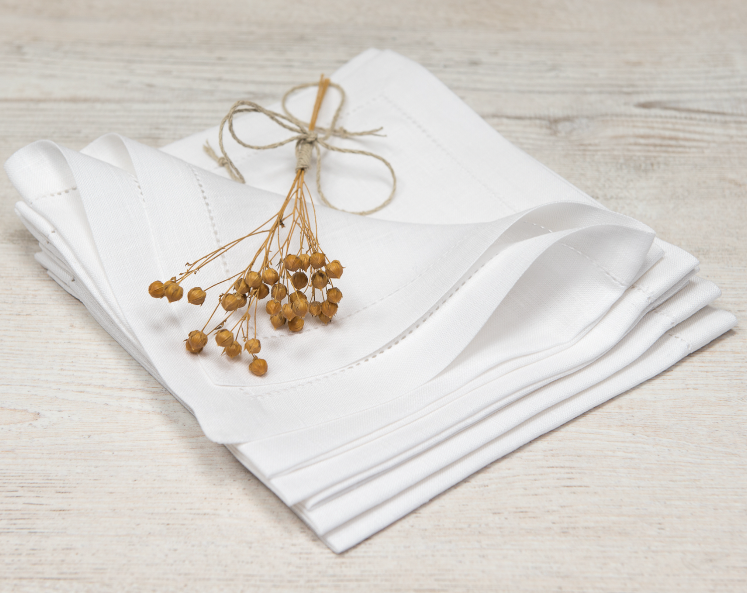 Flertal linneservetter i vit med hålsöm ligger vikta i en hög tillsammans med torkad lin. 