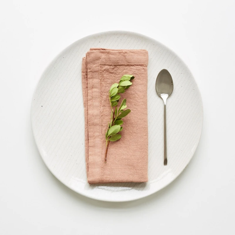 Handsydd creme färgad tygservett i inne som ligger vikt på en vit tallrik med en grön kvist.