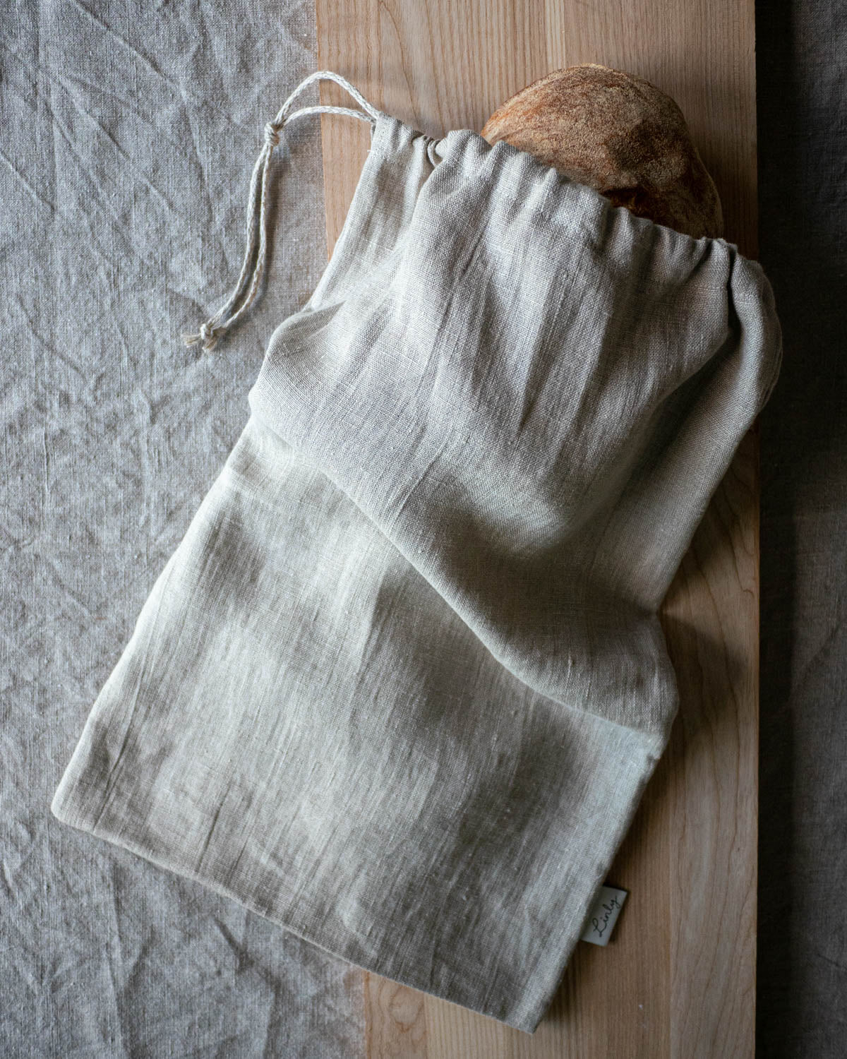 Brödpåse i linne ligger på en skärbräda på en linneduk. Påsen innehåller ett suredgsbröd som skymtar ut genom öppningen.