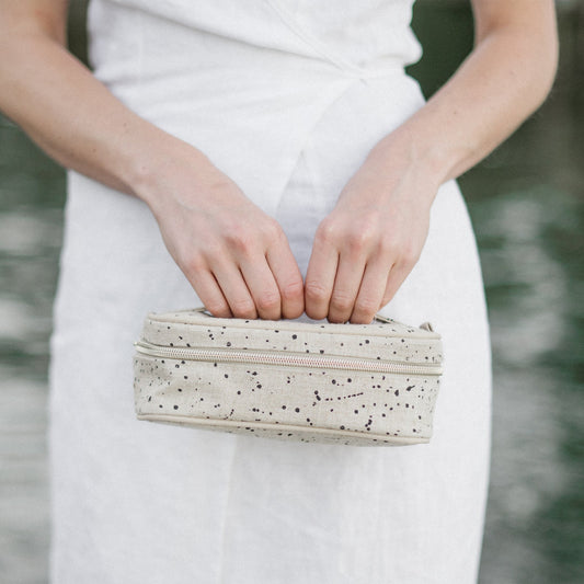 Kvinner håller i en liten rektangulär necessär i naturell linne med svarta prickar i olika storlekar.