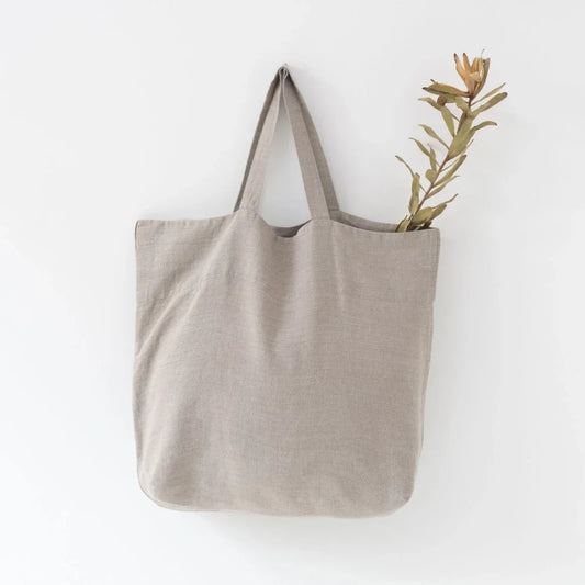Medelstor tygväska i naturell linne som hänger på en krok. Väskan har formen av en rektangel med medellånga handtag. Från väskan sticker en grön torkad kvist upp. 
