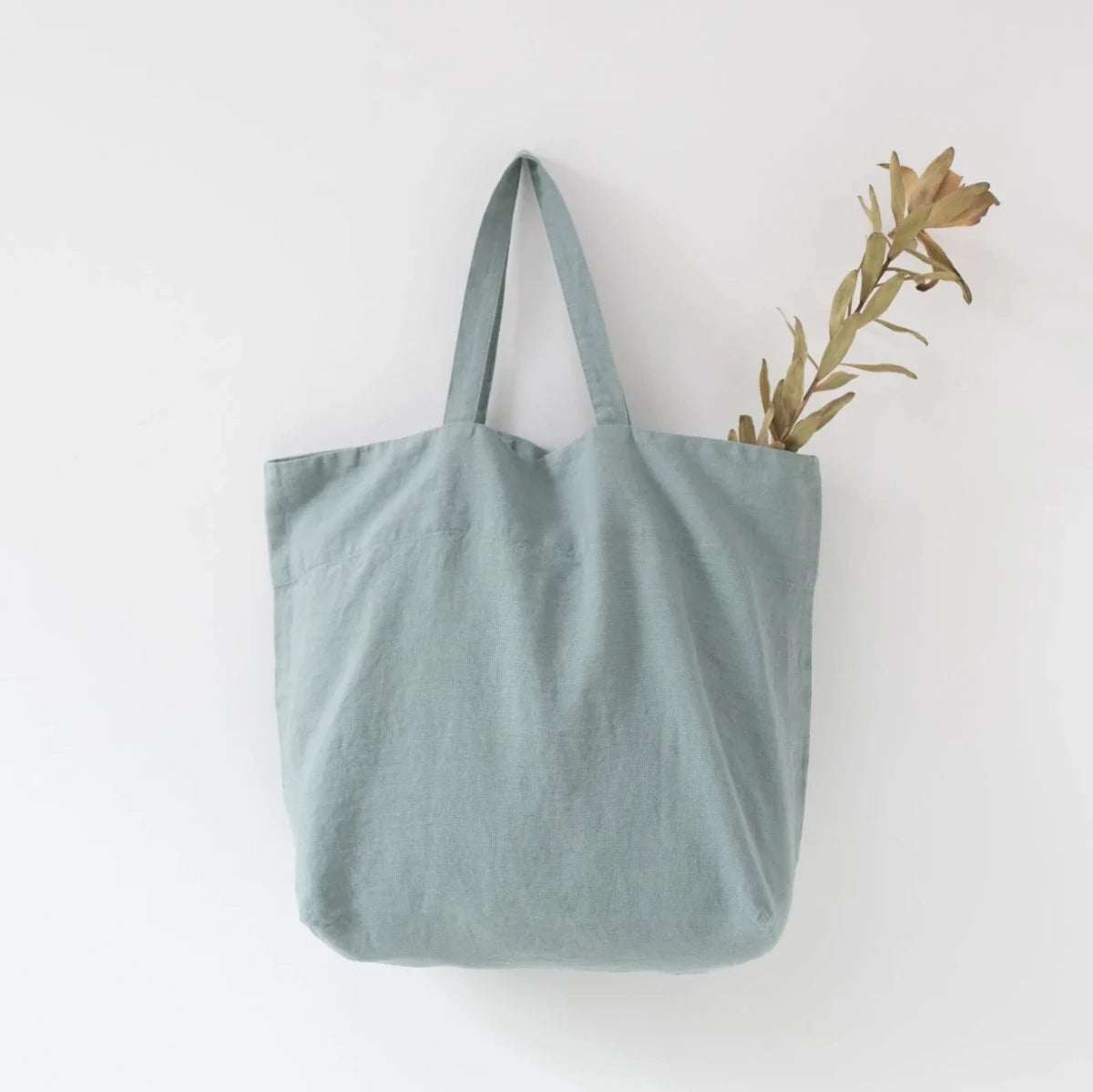 Medelstor tygväska i turkos linne som hänger på en krok. Väskan har formen av en rektangel med medellånga handtag. Från väskan sticker en grön torkad kvist upp.
