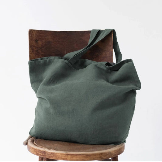 Medelstor tygväska i skogsgrön linne som står på en trästol. Väskan har formen av en rektangel med medellånga handtag. Väskan är välfylld och handtagen hänger över ena stolskanten