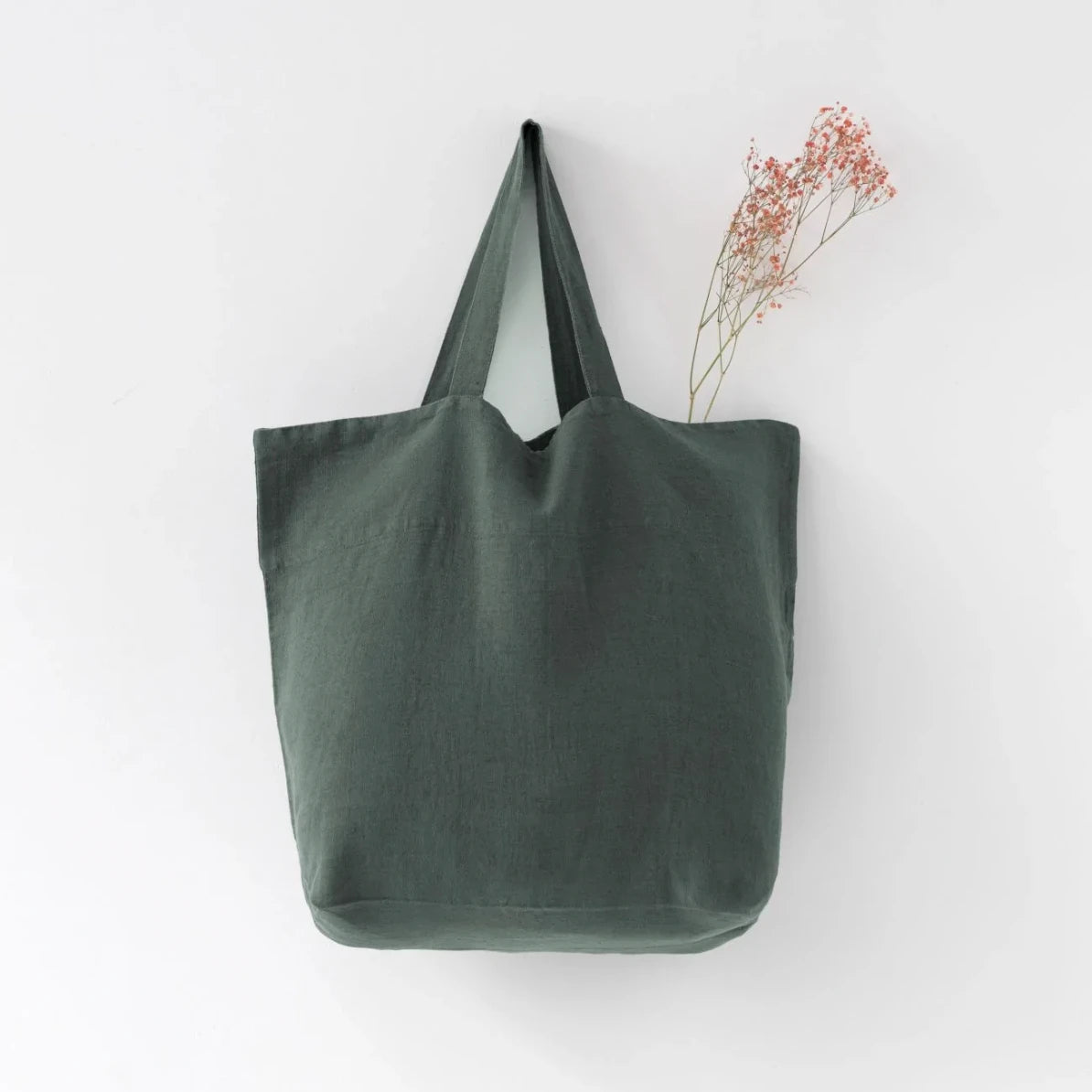 Medelstor tygväska i skogsgrön linne som hänger på en krok. Väskan har formen av en rektangel med medellånga handtag. Från väskan sticker en grön torkad kvist upp.