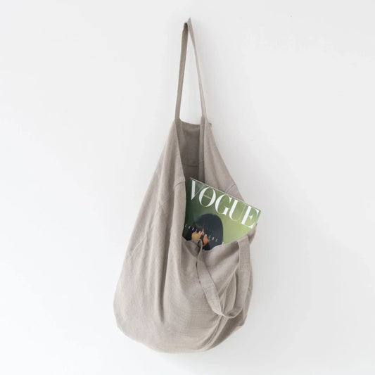 Medelstor tygväska i naturell linne som hänger på en krok med sitt ena handtag. Väskan har formen av en rektangel med medellånga handtag. Från väskan skymtar en Vouge tidning.