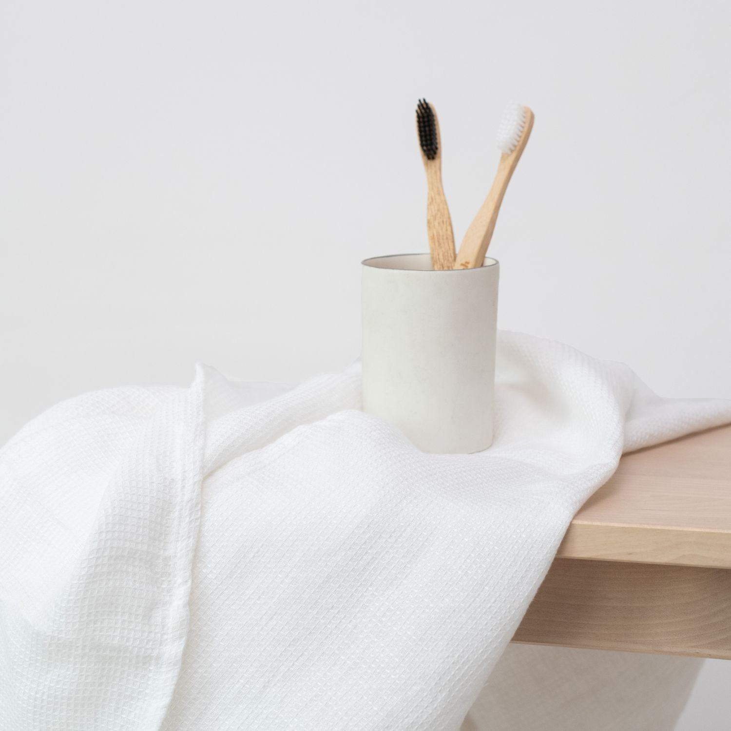 Liten våfflad handduk i vit som ligger över en träbänk. På den våfflade handduken står ett glas med tandborstar.