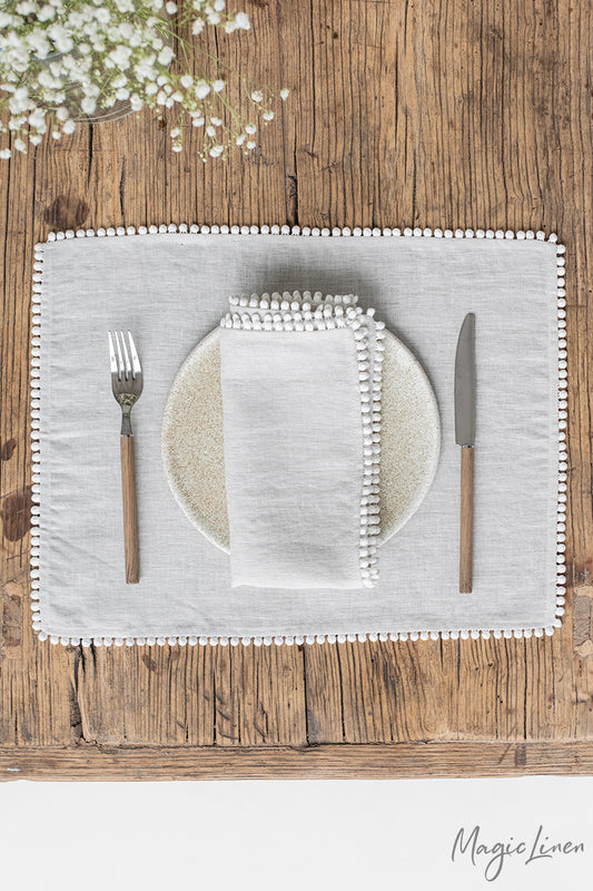 Rektangulär ljusgrå bordstablett i linne med en kantdetalj som är gjord av små tygbollar. Bordstabletten ligger på ett rustikt träbord med en vit tallrik och ljusgrå linneservett med kantdetalj. 