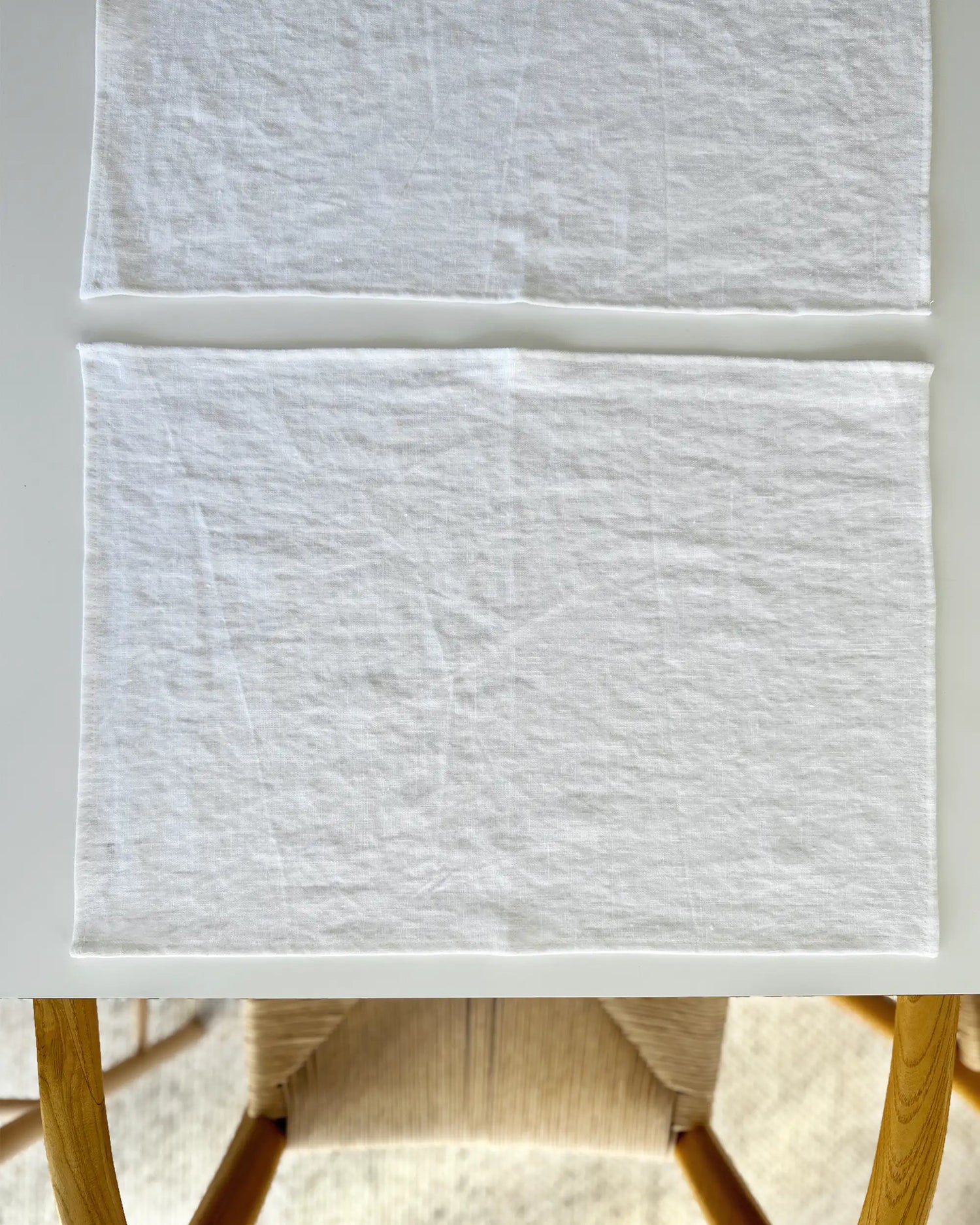 Två handsydda bordstabletter i vitt linnetyg som ligger på ett bord. 