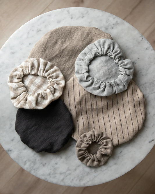 Flera handsydda skålöverdrag i 100% linne ligger på ett marmor bord. Alla skålöverdrag har olika storlekar och jordnära nyanser och mönster. 