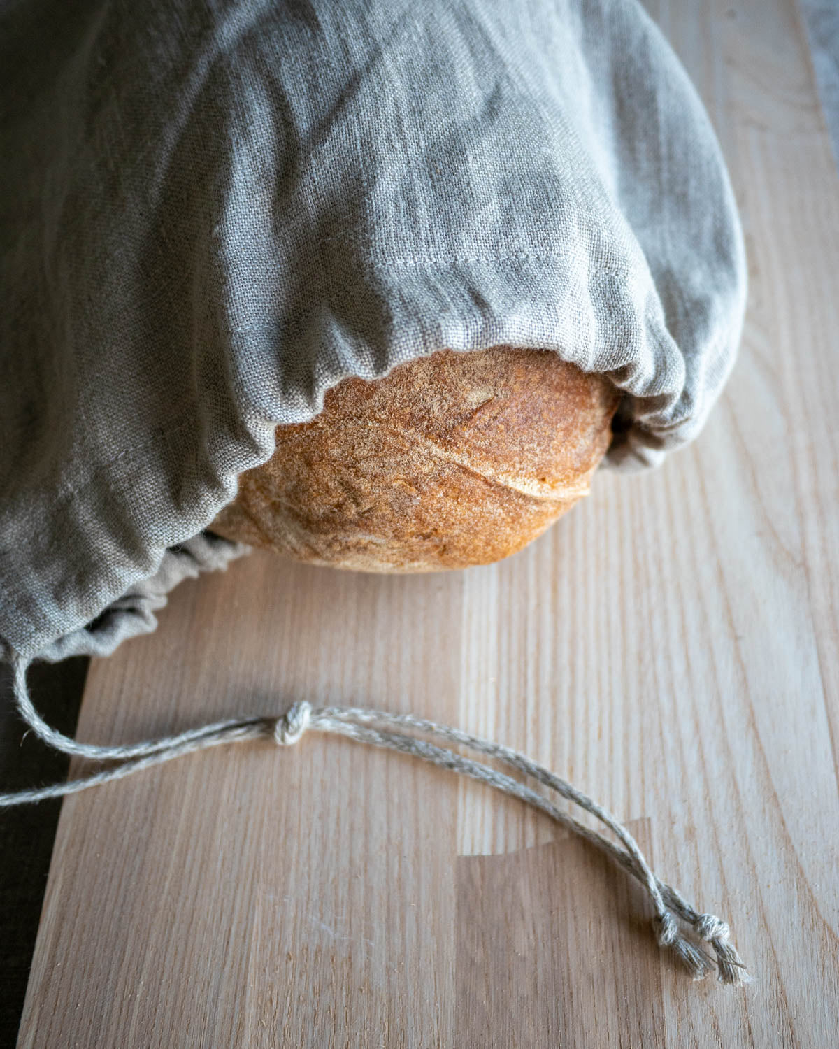 En brödpåse i naturell linnetyg som ligger på ett träbord. I påsen ligger ett bröd. Ett smalt linneband är fäst på påsen som du kan dra i för att stänga den.