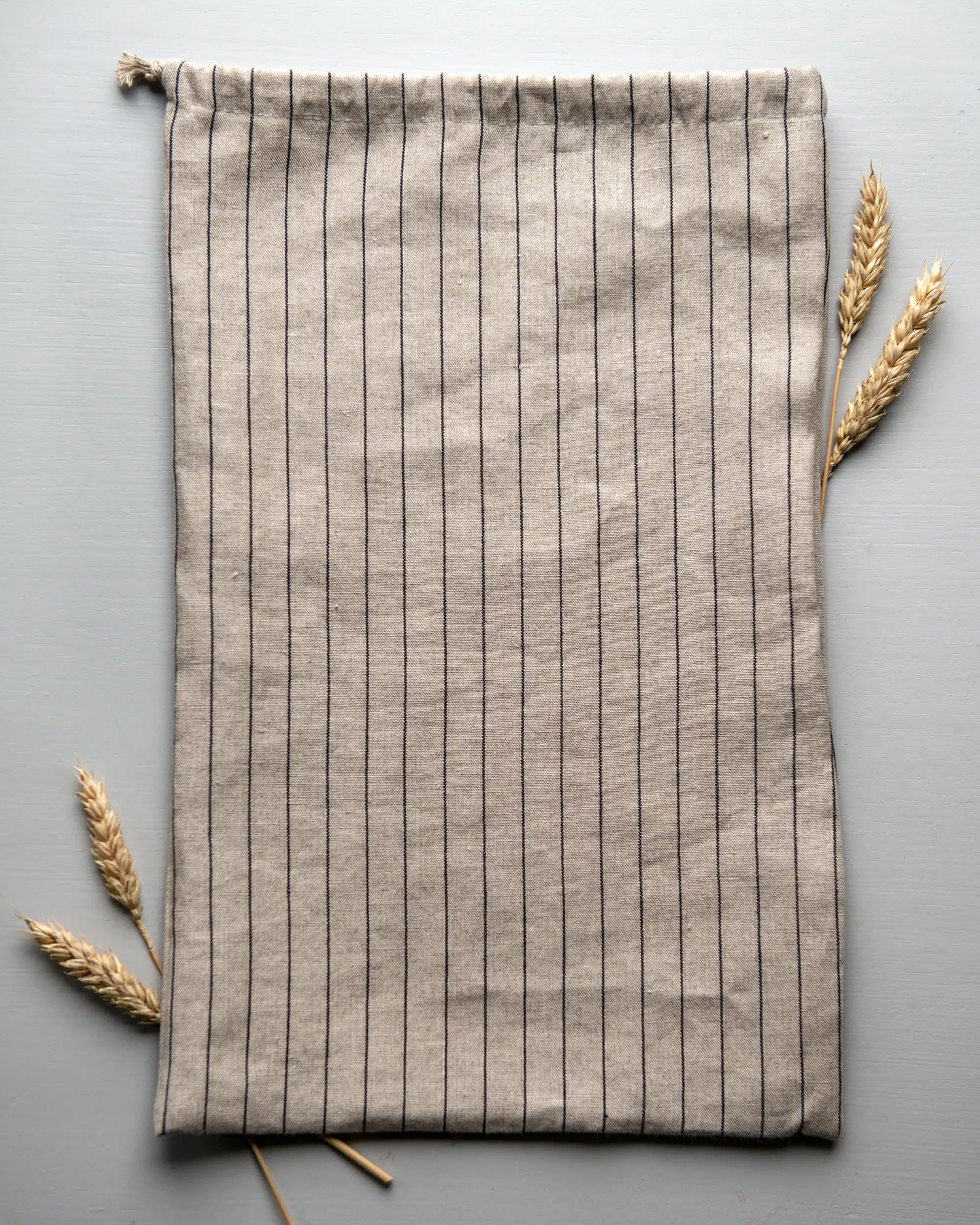 En handsydd brödpåse i randigt linnetyg som ligger på en yta tillsammans med torkat vete. 