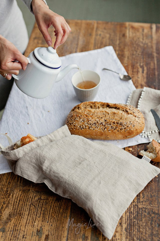 En rektangulär brödpåse i naturell linne som ligger på ett trä bord tillsammans med bröd  och tekanna. 