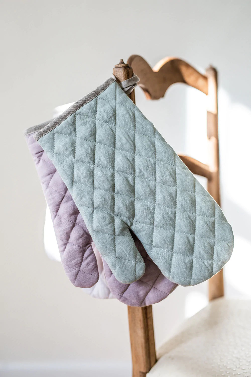 Två stycken quiltade grytvantar i linne hänger i sin ögla på en stolskant.