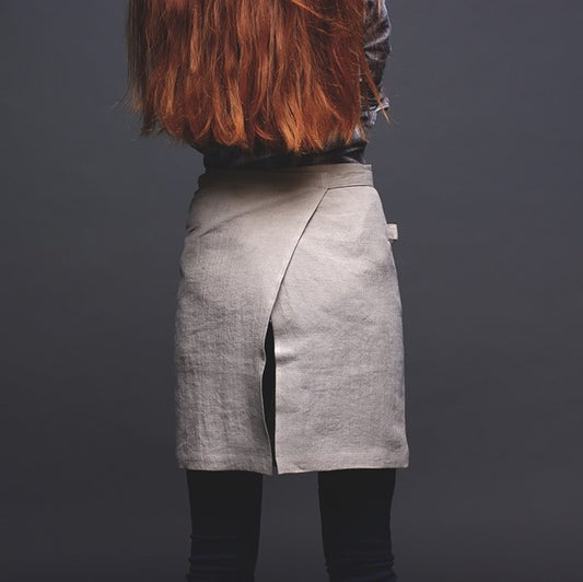 Ett mindre köksförkläde i naturell linne som bärs utav en kvinna. Kvinnan står med ryggen mot kameran och förklädet täcker bakdel och går omlott. 