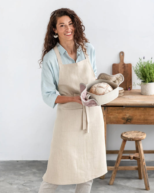 Kvinna med lockigt hår har på sig ett linneförkläde, ihållandes i två grytlappar som omsluter en varm form med nybakat bröd. 