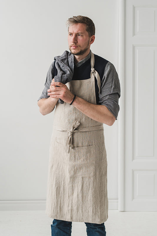 En man bär ett linneförkläde i beige som går ner till knäna. Mannen håller i en kökshandduk. 