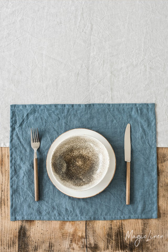 Rektangulär blå bordstablett i linne som ligger på ett rustikt träbord med en vit linneduk. På bordstabletten står det en vit tallrik med bestick.