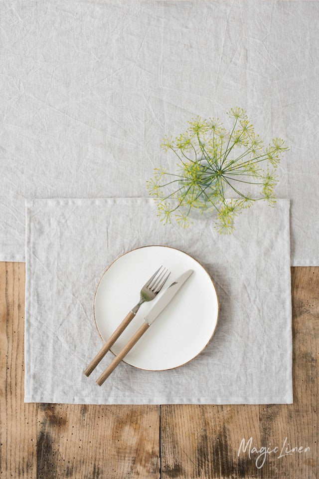 Rektangulär bordstablett i ljusgrå linne som ligger på ett rustikt träbord med linneduk. På bordstabletten står det en liten vit tallrik med bestick.