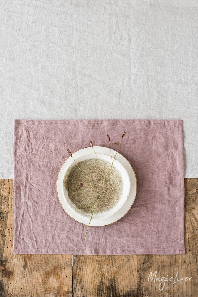 Rektangulär ljusrosa bordstablett i linne som ligger på ett rustikt träbord med en vit linneduk. 