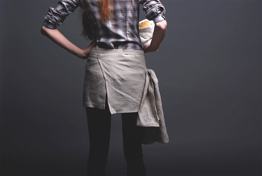 En kvinna bär ett mindre köksförkläde i naturell linne och står med ryggen mot kameran. Köksförklädet går omlott och täcker ovanlåren.