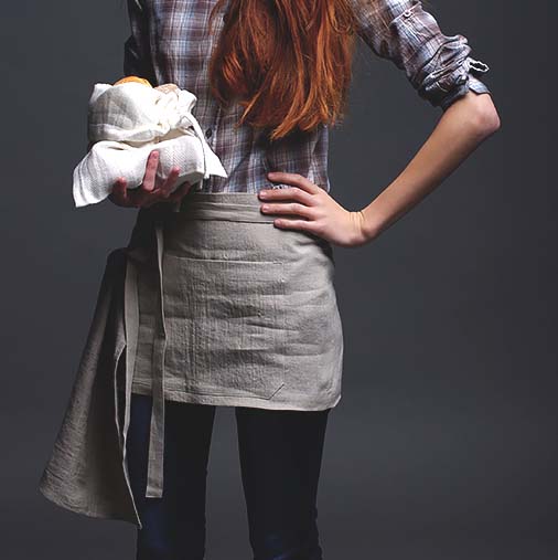 En kvinna har på sig ett miljövänligt köksförkläde i naturell linne. Köksförklädet har knutits i midjan och täcker ovanlåren.