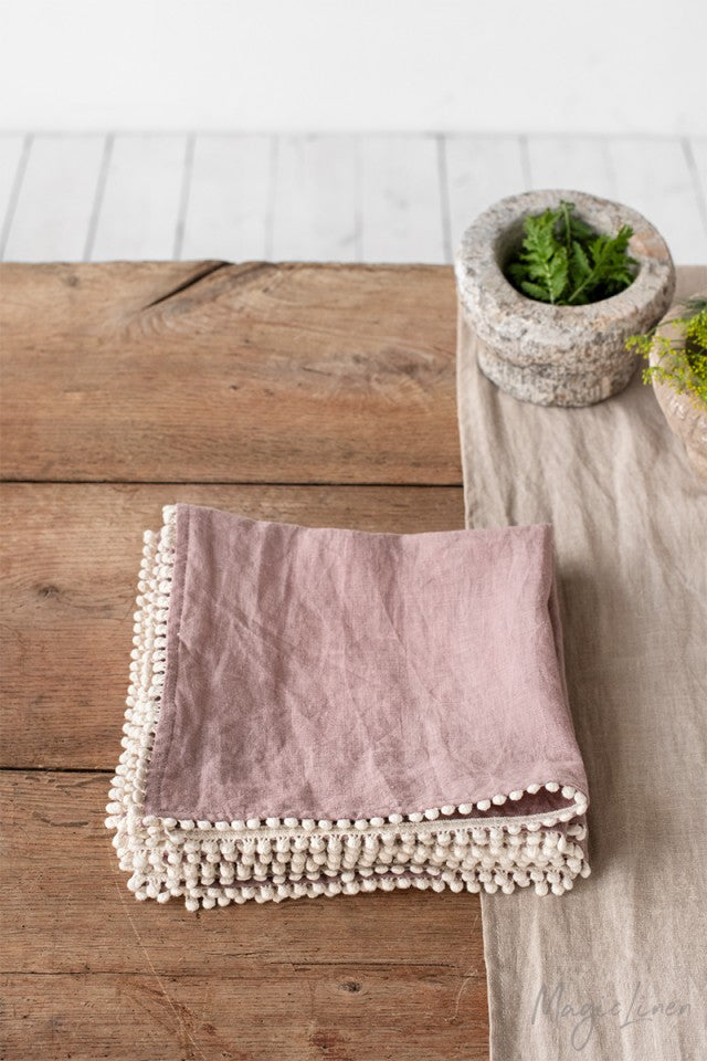 Ljusrosa linneservetter med en kantdetalj i form av små dekorativa tygbollar. Servetterna ligger vikta i en hög på ett rustik träbord med en ljusgrå linneduk. På bordet står en mortel med kryddor i.