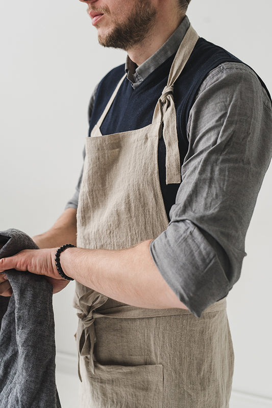 Köksförkläde i naturellt linne som bärs utav en man. Förklädets band har knutits runt magen och har träts över huvudet. 
