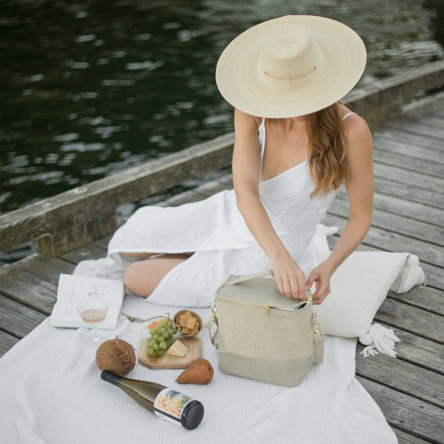 En kvinna sitter på en brygga och har picknick och öppnar en kylväska i naturell linne med grön kant.